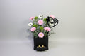 Floral arrangement with pink flowers and greenery, a black box with gold candies, and a 'mom' keychain on a light gray background.