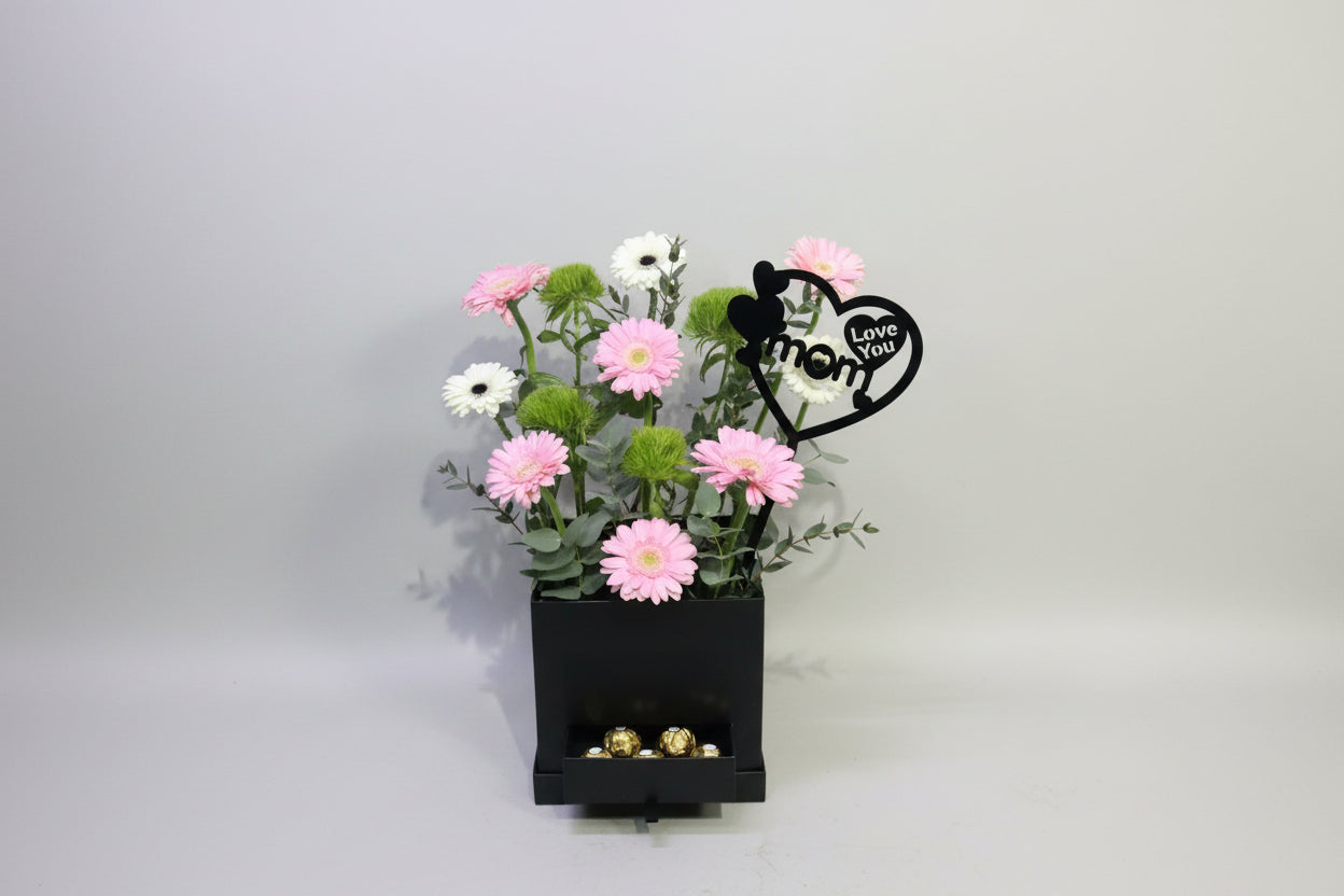 Floral arrangement with pink flowers and greenery, a black box with gold candies, and a 'mom' keychain on a light gray background.